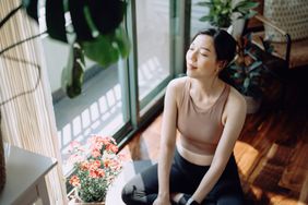 Active young Asian sports woman taking a break after working out at home, sitting on exercise mat taking a deep breath with her eyes closed. Sports and exercise routine. Health, fitness and wellness concept