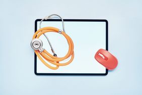 Top view of tablet computer with orange colored stethoscope and red computer mouse on top, all against a light blue background