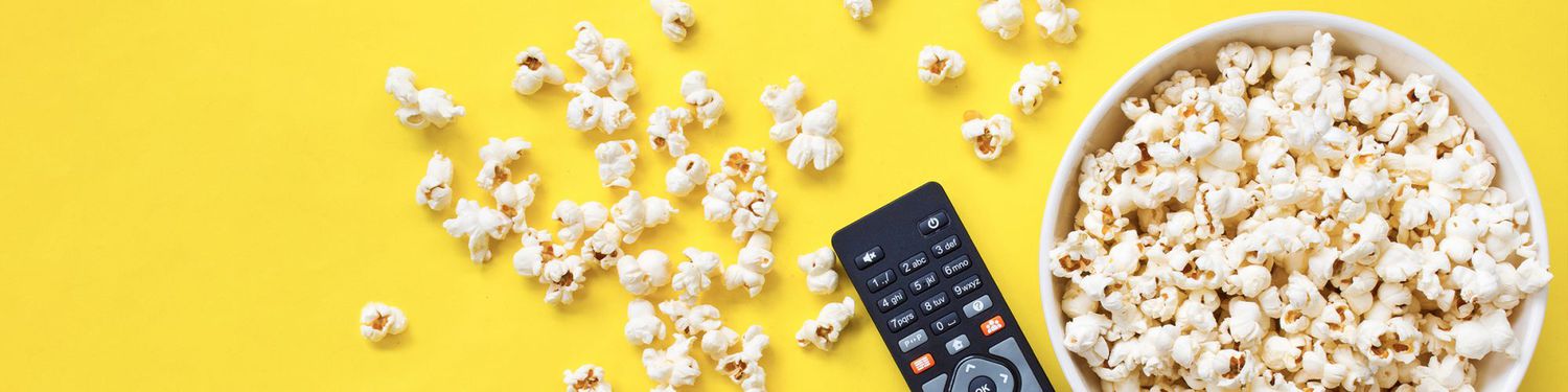 Black TV remote next to popcorn in a white bowl, some spilled out, all on a yellow surface 