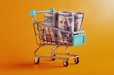 Shopping cart with rolled American One Hundred Dollar Bills on yellow / orange background