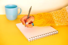 Woman in yellow white knitted sweater with bright multi colored manicure is writing in notebook sitting at yellow table. Light blue cup stands next to her hand.